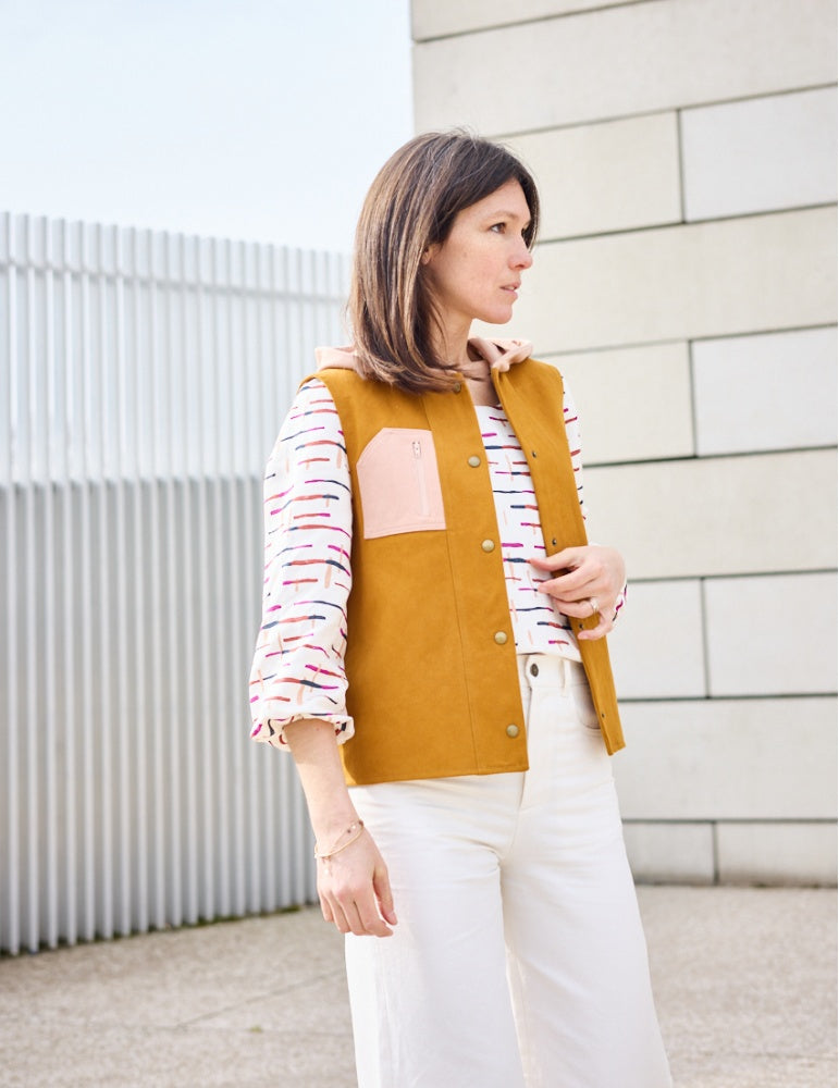 Woman wearing the Alcove Vest sewing pattern from Atelier Scämmit on The Fold Line. A sleeveless jacket pattern made in wools or gabardine fabrics, featuring a full lining, hip length, hood, button and snap closure, and zipped upper pocket.