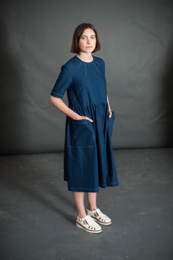 Woman wearing the Ellis Dress sewing pattern by Merchant and Mills. A dress pattern made in linen, light wools, and light to medium weight cotton fabric featuring four delicate neck darts, set in elbow length sleeves, gathered waist and bold patch pockets.