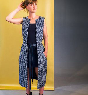 Woman wearing the Aestiva Sleeveless Vest sewing pattern from Wearologie on The Fold Line. A sleeveless vest pattern made in wool, satin, chambray, gabardine or jacquard fabrics, featuring gathers shaping the waist, midi length, wide round neckline, pockets highlighted with piping and a front fastening using a belt.