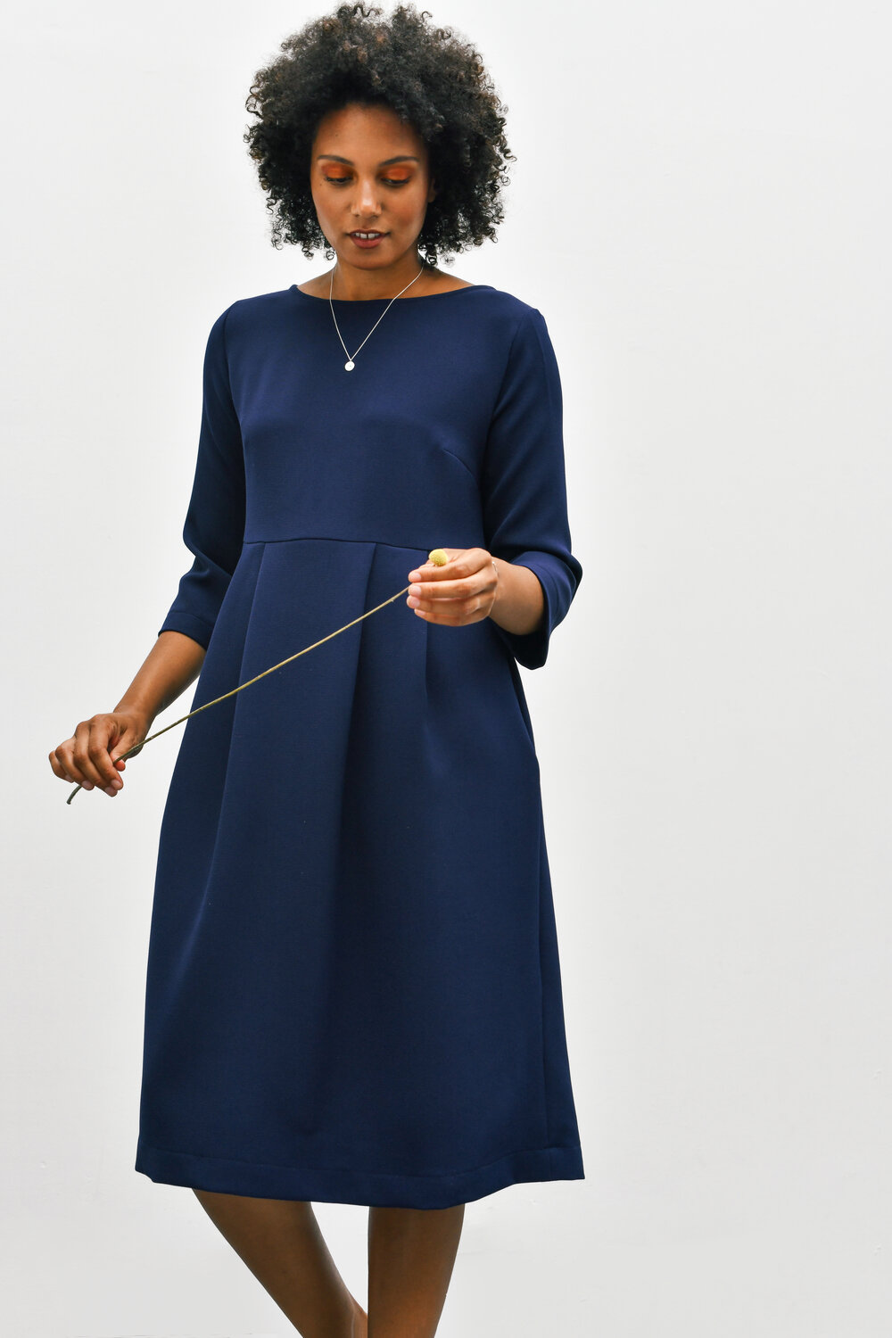 Woman wearing the Cissy Dress sewing pattern by Homer and Howells. A dress pattern made in cottons, cords, wool or crepe fabrics, featuring a slightly boxy fit, boat neck, bracelet length sleeves, centre back zip. Deep pleats in the skirt create volume and movement, plus side seam pockets.
