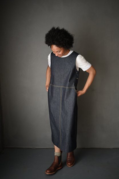 Woman wearing the Whittaker Dress sewing pattern by Merchant and Mills. A dress pattern made in denim, cotton twill, corduroy or moleskin fabric featuring a classic pinafore style, button side fastening, side and back pockets plus topstitching.