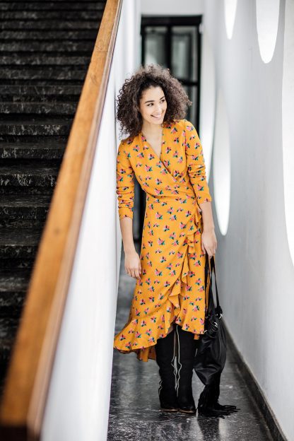 Woman wearing the Charlotte Dress sewing pattern from Fibre Mood on The Fold Line. A wrap dress pattern made in crepe, linen, satin, Tencel or muslin fabrics, featuring a deep V-neck, midi length with high low ruffle hem, waist tie, and 3/4 sleeve.