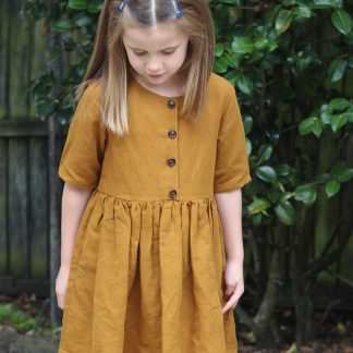 Child wearing the Baby/Child Kauri Dress sewing pattern by Below the Kowhai. A dress pattern made in light to medium weight linen, cotton, flannel or sateen fabrics, featuring a gathered waist, relaxed fit, pockets, elbow length sleeves and button bodice.