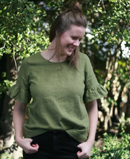 Woman wearing the Rātā Top sewing pattern by Below the Kowhai. A blouse pattern made in light to medium weight cotton, chambray, poly cotton, linen, rayon, sateen or tencel fabrics, featuring a round neckline, button and loop back closure and a sleeve ruffle.