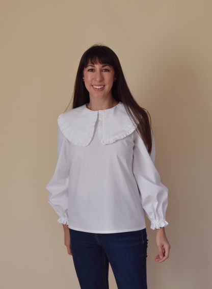 Woman wearing the Bakerloo Blouse sewing pattern from Nina Lee on The Fold Line. A blouse pattern made in lawn, poplin, chambray, silk dupion, crepe or fine needlecord fabrics, featuring a large ruffled collar, balloon sleeves with elasticated cuffs and ruffled edges, button and keyhole closure at the centre-back and loose fitting around the waist.