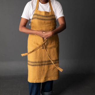 Woman wearing the Unisex Workday Apron sewing pattern by Merchant and Mills. A long length apron pattern made in cotton canvas, cotton twill, medium to heavy weight linen or oilskin fabric featuring three pockets, waist ties and adjustable neck strap.