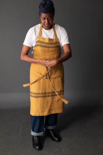 Woman wearing the Unisex Workday Apron sewing pattern by Merchant and Mills. A long length apron pattern made in cotton canvas, cotton twill, medium to heavy weight linen or oilskin fabric featuring three pockets, waist ties and adjustable neck strap.