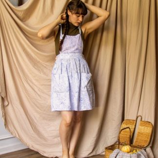 Woman wearing the Fleur Pinafore sewing pattern from Untitled Thoughts on The Fold Line. A pinafore pattern made in linen, chambray, cotton poplin, rayon/linen blend, quilting cotton, mid- twill or light to mid-weight corduroy fabrics, featuring tie shoulder straps, patch pockets, bib, deep waistband with gathered skirt and side button closure.