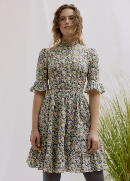 Woman wearing the Alexa Frill Dress sewing pattern by Liberty Sewing Patterns. A dress pattern made in cotton, satin or poplin fabrics, featuring elbow length sleeves with frill, stand frill collar, fitted bodice, flared skirt with tiered frill and back zipper closure.