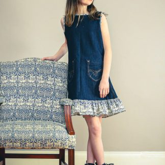 Child wearing the Jeans to Child/Teen Dornoch Dress sewing pattern by Greyfriars and Grace. A sleeveless dress pattern made in pre-loved denim fabrics, featuring an A-line silhouette, front pockets, back zip closure, hem ruffle, and contrasting bias binding on the neckline and armholes.