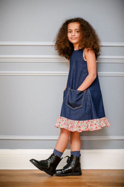 Child wearing the Jeans to Children's Joppa Dress sewing pattern by Greyfriars and Grace. A sleeveless dress pattern made in pre-loved denim fabrics, featuring an A-line silhouette, front pockets, hem ruffle and contrasting bias binding on the neckline, back neck tie and armholes.
