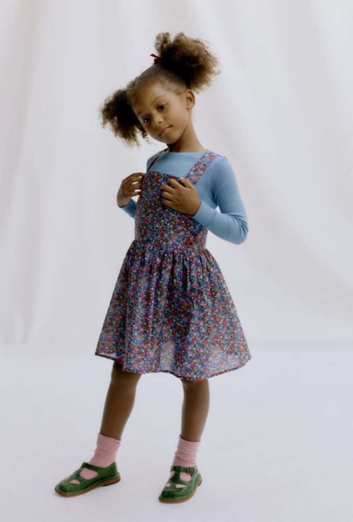 Child wearing the Child/Teen Kiki Dungaree Set sewing pattern by Liberty Sewing Patterns. A pinafore pattern made in denim, corduroy or cotton fabrics, featuring a zip side opening, button fastening straps, flared skirt and knee length finish.
