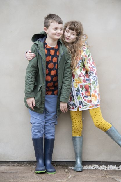 Children wearing the David Raincoat sewing pattern from Bobbins and Buttons on The Fold Line. A unisex raincoat pattern made in laminated cotton, polyurethane, coated cotton, cotton drill or denim fabrics, featuring a zip-front, bound edges, raglan sleeves, panelled hood, big boxy pockets or pockets with flaps.