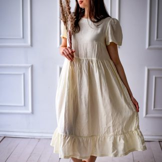 Woman wearing the Naya Dress sewing pattern from Kates Sewing Patterns on The Fold Line. A dress pattern made in cotton or linen fabrics, featuring a high waistline, bust darts, voluminous sleeves with gathers at the top, full skirt gathered at the waist with hem ruffle, closure with button/hook at back neckline and midi length hem.