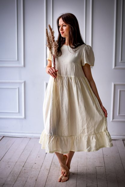 Woman wearing the Naya Dress sewing pattern from Kates Sewing Patterns on The Fold Line. A dress pattern made in cotton or linen fabrics, featuring a high waistline, bust darts, voluminous sleeves with gathers at the top, full skirt gathered at the waist with hem ruffle, closure with button/hook at back neckline and midi length hem.