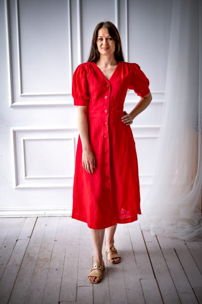 Woman wearing the Stella Dress sewing pattern from Kates Sewing Patterns on The Fold Line. A dress pattern made in cotton, linen or silk fabrics, featuring shoulder gathers, short voluminous sleeves with gathers at the top and bottom, below knee length flared skirt, side pockets, front button fastening and a belt comes out of the waist darts and ties at the back.