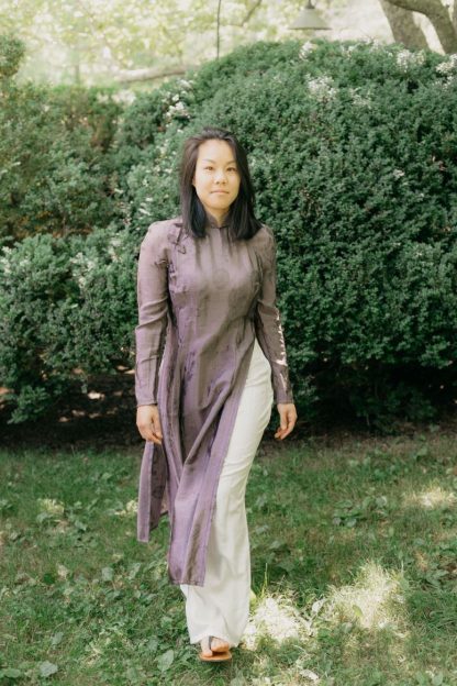 Woman wearing the 139 Vietnamese Áo Dài sewing pattern from Folkwear on The Fold Line. A tunic and trouser pattern made in brocade, silk, polyester or cotton gauze fabrics, featuring a tunic with front and back waist shaping darts, band collar, front neckline/armhole opening, tapered long raglan sleeves, long free-hanging front and back panels. The wide legged trousers have a left side zipper and faced waistline.