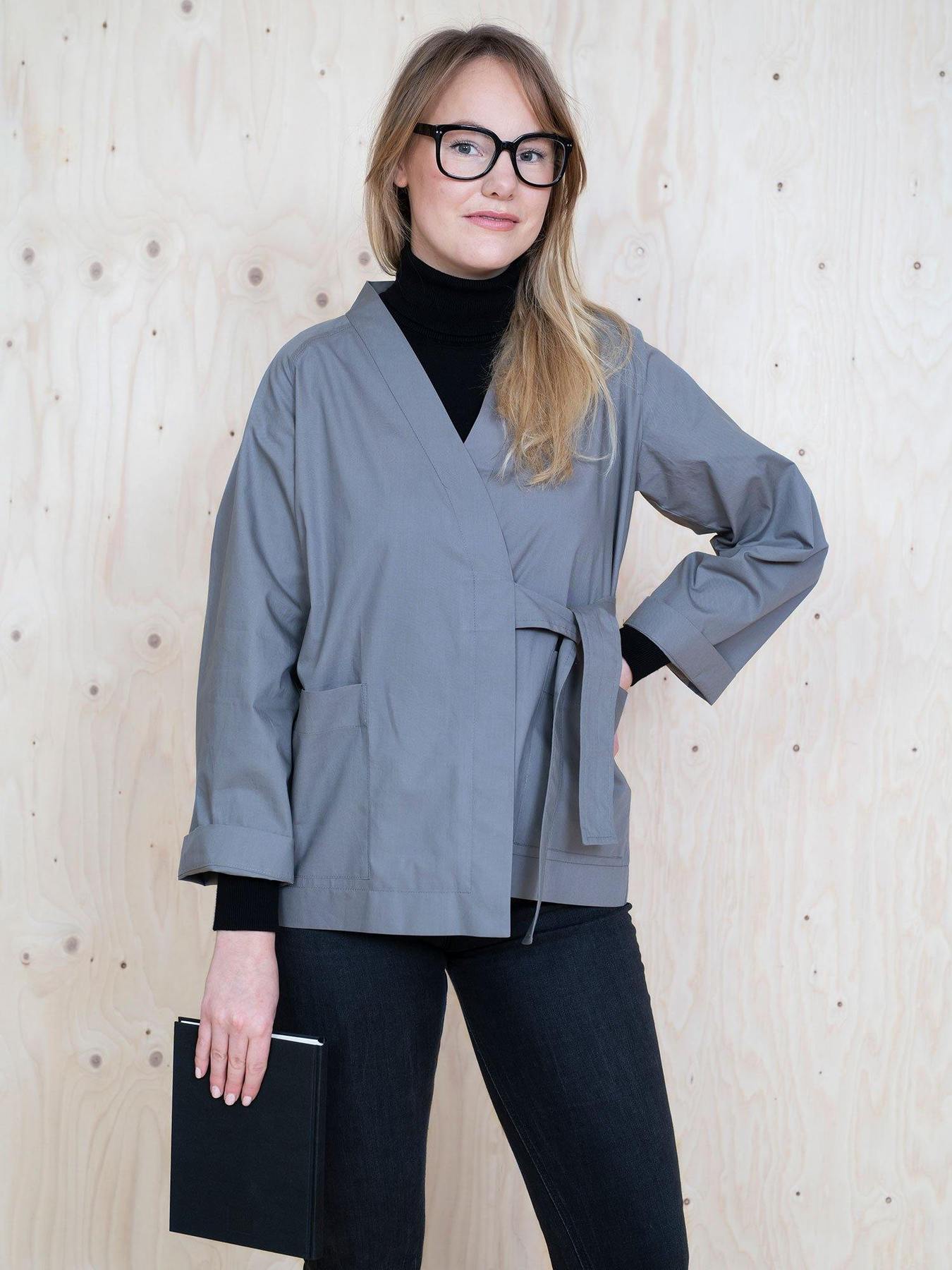 Woman wearing the Wrap Jacket sewing pattern from The Assembly Line on The Fold Line. A jacket pattern made in cotton twill, denim or lightweight canvas fabrics, featuring large pockets, full length sleeves turned back, crossover front with matching fabric tie and relaxed fit.