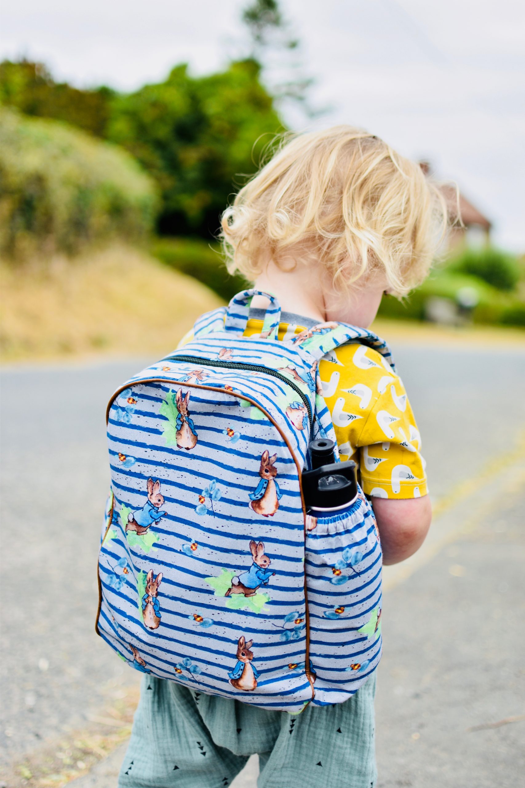Child wearing the Children's Back to Cool Backpack sewing pattern from Waves & Wild on The Fold Line. A rucksack pattern made in canvas, twill or denim fabrics, featuring side pockets, adjustable wide shoulder straps, zip closure and piping.