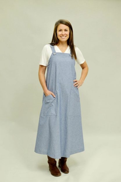 Woman wearing the Pinafore Dress sewing pattern from Folkwear on The Fold Line. A pinafore pattern made in cotton broadcloth, chambray, poplin, linen, wool, sturdy knits, wool, denim, twill, or canvas fabrics, featuring a low-calf-length, two front patch pockets with pocket bands, wide shoulder straps that button in front, and a relaxed fit.