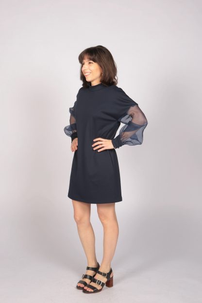 Woman wearing the Audrey Dress sewing pattern from Camimade on The Fold Line. A dress pattern made in tartan, gabardine, velvet, tweed, denim, jacquard, silk, woollen crepe, triple crepe or heavy jersey fabrics, featuring an above knee length, loose fit, grown-on long puff sleeves, high collar, back invisible zip, small buttons on the collar and pockets.