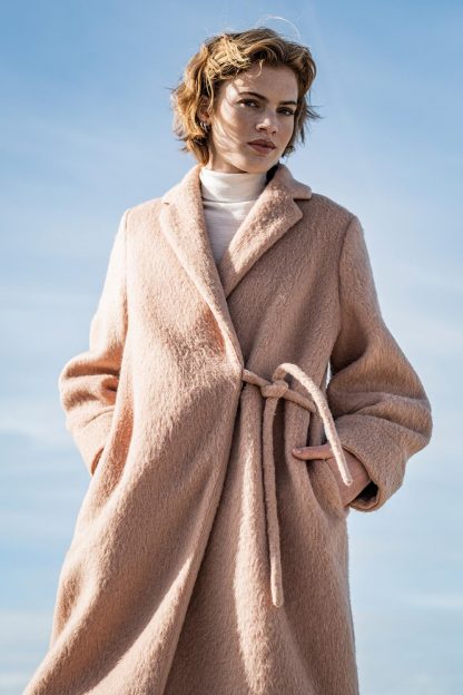 Woman wearing the Carmen Coat sewing pattern from Fibre Mood on The Fold Line. A coat pattern made in wools, boucle, plush or long-haired faux fur fabrics, featuring collar and lapels, shaped sleeves, in-seam pockets, slight cocoon shape, left sided tie belt and knee length finish.