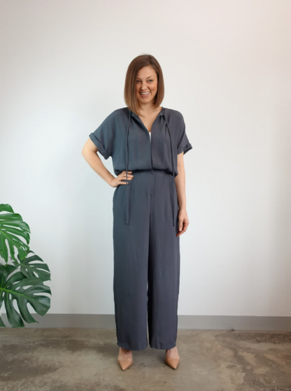 Woman wearing the Eadie Woven Jumpsuit Dress sewing pattern from Style Arc on The Fold Line. A jumpsuit pattern made in silk, crepe or rayon fabrics, featuring an extended shoulder line, turned up sleeve cuffs, elastic waist, front opening, snap closure and bound neck with ties and wide legs.
