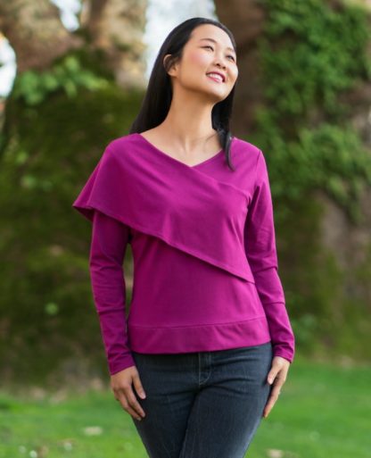 Woman wearing the Carmo Top sewing pattern from Itch to Stitch on The Fold Line. A knit top pattern made in jersey, sweater knit or French terry fabrics, featuring an asymmetrical silhouette, front and back V-necklines, diagonal scarf which wraps over one shoulder and around the opposite side, long sleeves finished with cuff bands and bodice is finished with a bottom band.