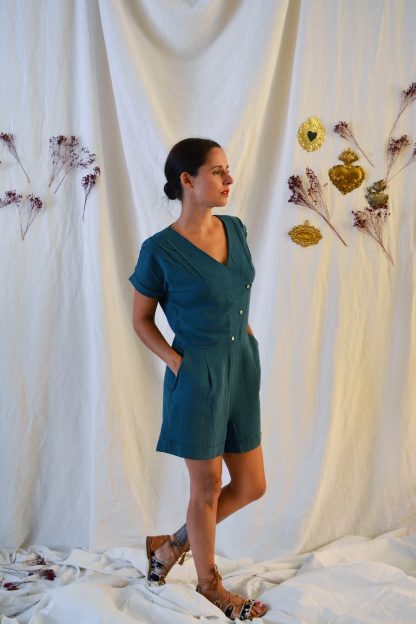 Woman wearing the Eclipse Playsuit and Dress sewing pattern from Maison Fauve on The Fold Line. A playsuit pattern made in cotton poplin, tencel, light denim, viscose, crepe or cotton satin fabrics, featuring angled front pockets, V-neck, short sleeves with turn-ups, shoulder pleats, 3 button diagonal front fastening and turned up hems.