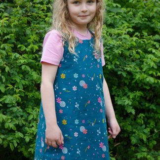Child wearing the Baby/Child Rosie Dress sewing pattern from Bobbins and Buttons on The Fold Line. A pinafore dress pattern made in cottons, lightweight denim, broadcloth, corduroy, needlecord or denim fabrics, featuring button through straps, round neck and contrast staggered hem.