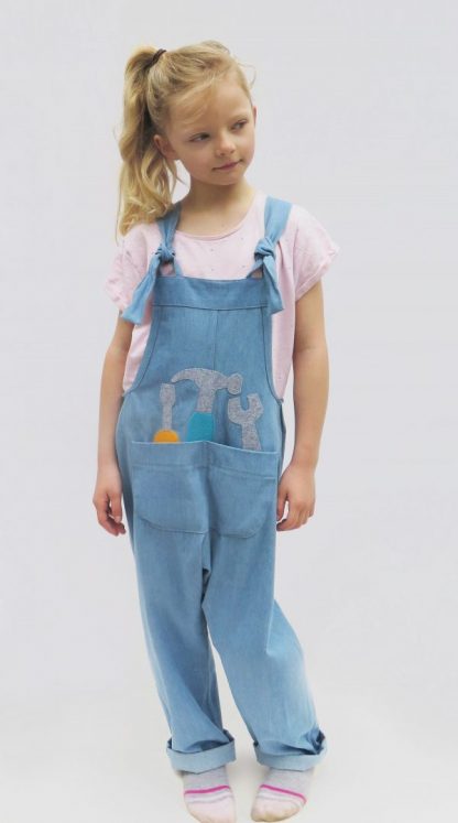 Child wearing the Children's Playtime Dungarees sewing pattern from Ruth Maddock Makes on The Fold Line. A dungarees pattern made in denim, canvas or corduroy fabrics, featuring elasticated shoulder straps with knot ties, large front pocket, applique options, relaxed fit, side pleats and outside seam finishes.
