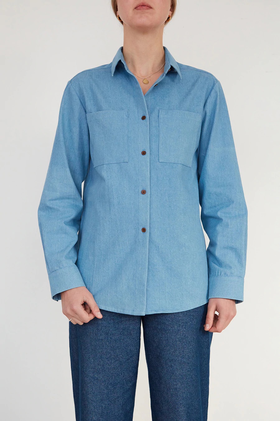 Woman wearing the Unisex Shirt sewing pattern from The Modern Sewing Co on The Fold Line. A shirt pattern made in poplin, lightweight twill or denim fabrics, featuring a boxy silhouette, collar and stand, front button closure, chest patch pockets, full length sleeves with button cuffs, back yoke with pleat and tab and high/low hem.
