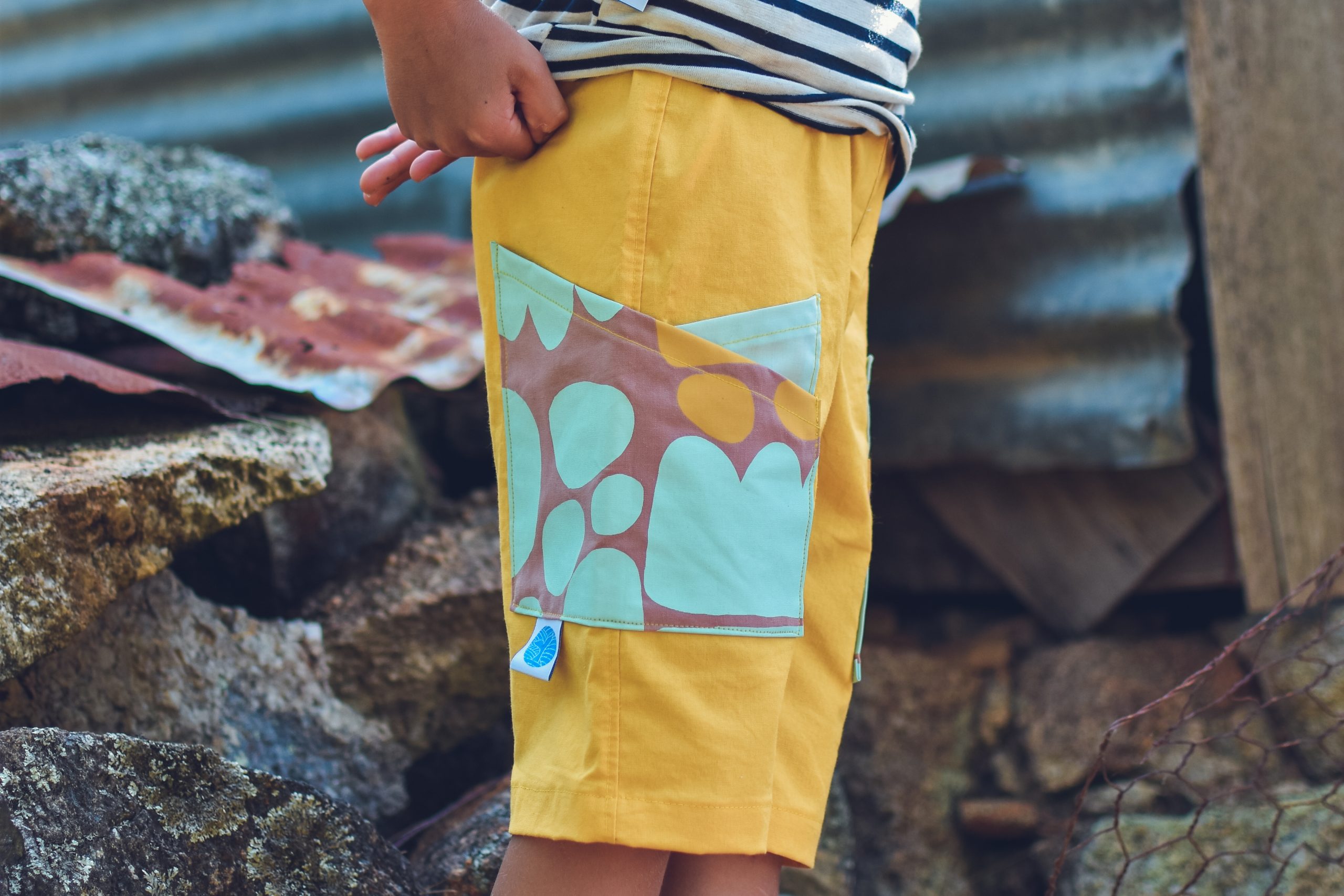 Child wearing the Baby/Child Crosswalk Shorts sewing pattern from Waves & Wild on The Fold Line. A shorts pattern made in denim or corduroy fabrics, featuring a crossover side pockets, soft elastic waistband, relaxed fit and knee length hem.
