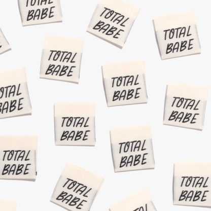 Photo showing 'Total Babe' Woven Labels from Kylie & The Machine on The Fold Line. A washable, durable and non-scratchy fabric label featuring a cream background, and the word 'Total Babe' in black text all ready to be sewn into your handmade clothes.