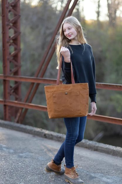 Woman holding the Pepin Tote sewing pattern from Noodlehead on The Fold Line. A tote pattern made in canvas or waxed canvas fabrics, featuring a front pocket with snap closure, divided interior pocket, recessed zip closure and shoulder length straps.