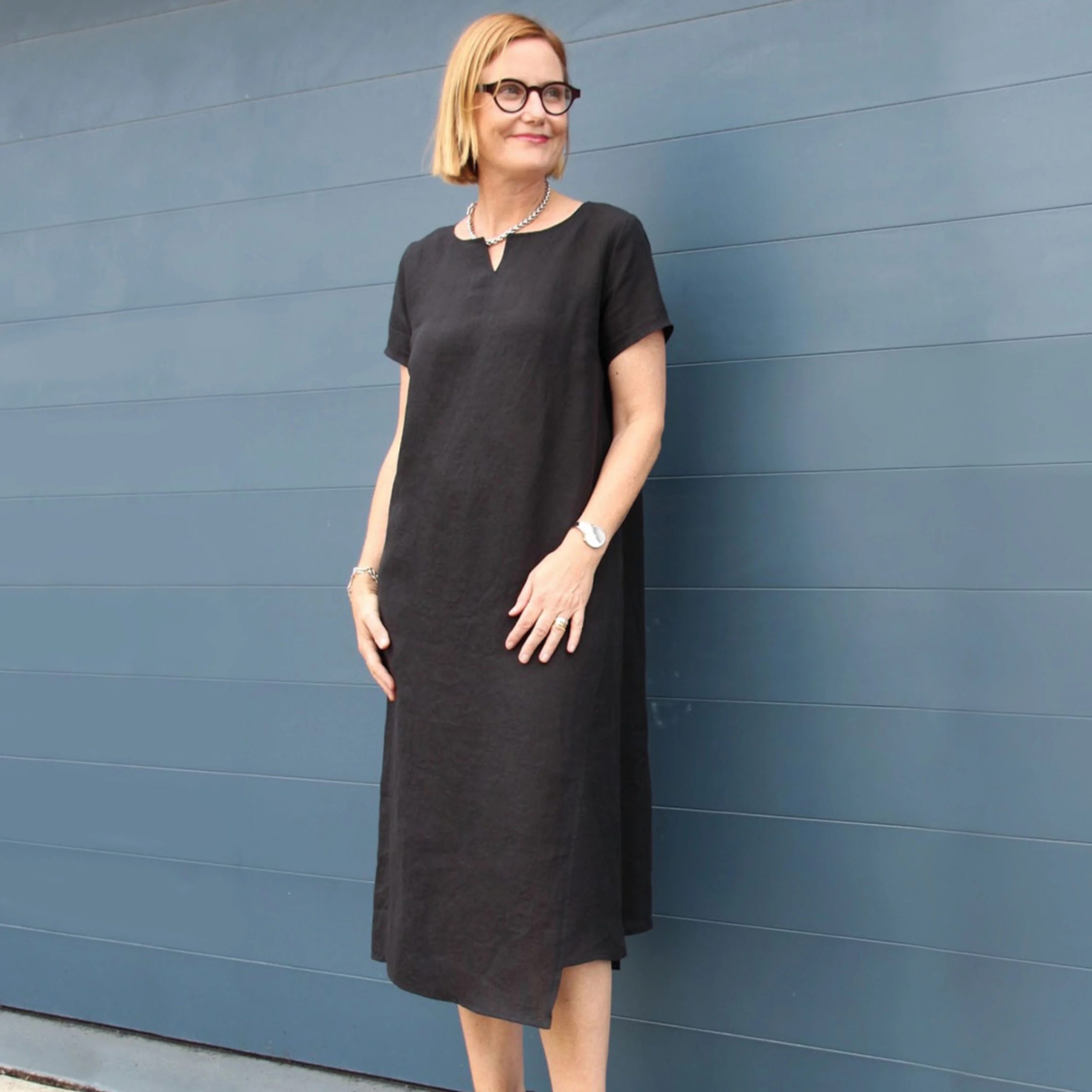 Woman wearing the Jane Dress sewing pattern from Tessuti Fabrics on The Fold Line. A dress pattern made in linen, linen/cotton, linen/viscose, medium weight drapey silk or lightweight wool crepe fabrics, featuring a round neckline with a small split, short sleeves, side pocket, side godet panel and asymmetrical midi length hem.