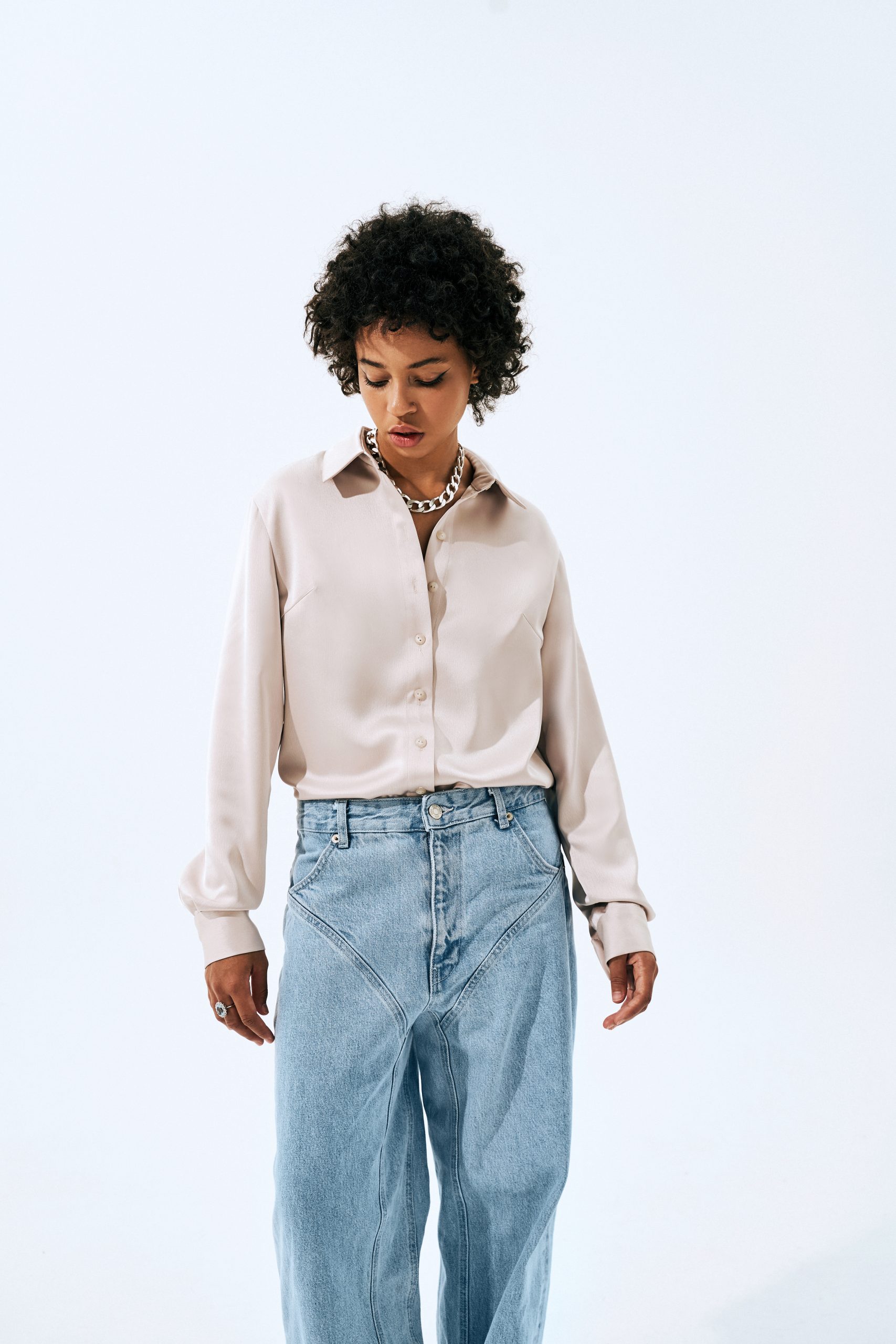 Woman wearing the Kaia Blouse sewing pattern from Vikisews on The Fold Line. A blouse pattern made in silk, artificial silk, cotton, linen, cupro, shirting, lightweight fine wale corduroy or modal fabrics, featuring a semi-fit, straight silhouette, bust darts, double-layer back yoke, turn-down shirt collar with separate collar stand, long set-in drop-shoulder sleeves, cuffs with button closure, front button closure and curved hem with side slits.