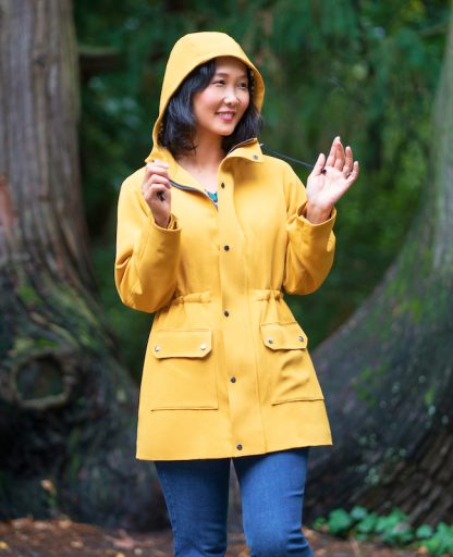 Woman wearing the Winterthur Jacket sewing pattern from Itch to Stitch on The Fold Line. A lined jacket pattern made in Twill, broadcloth, chambray, taslan nylon or linen fabrics, featuring front princess seams, back yoke, drawstring waist, front patch pockets, front zipper with buttoned flap, three-piece hood, long sleeves with snap closure and mid-thigh length.