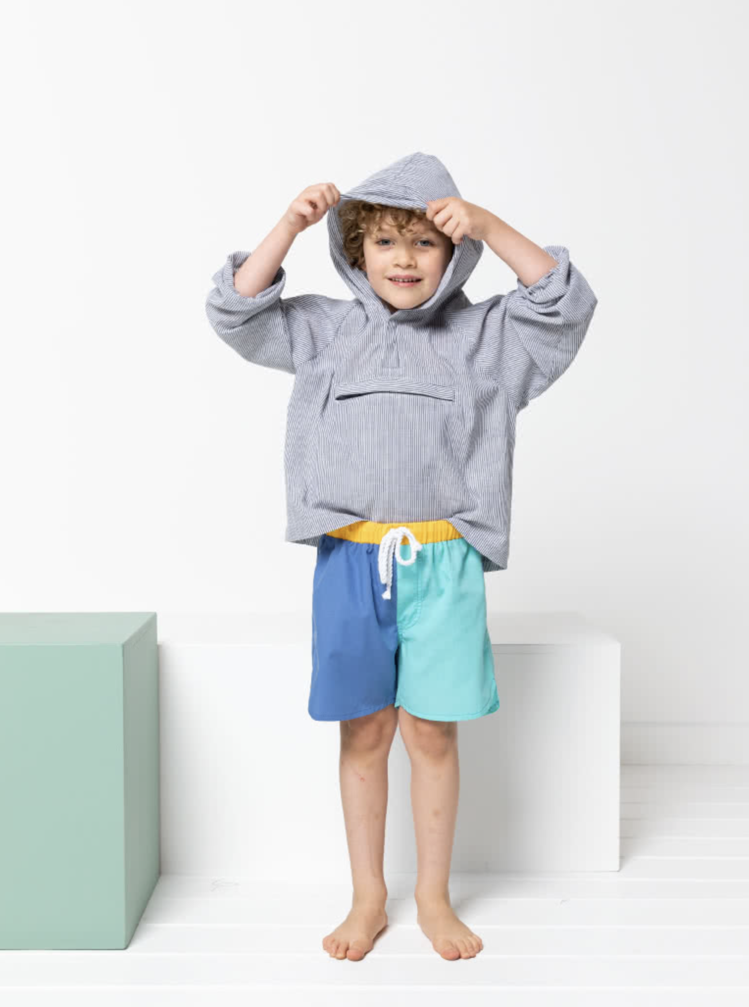 Child wearing the Children's Ash Anorak sewing pattern from Style Arc on The Fold Line. An Anorak pattern made in showerproof nylon fabric, cotton, or polyester fabrics, featuring a hip-length, unlined hood, raglan sleeves, elasticated cuffs, front neck tab snaps and centre front zip pocket.
