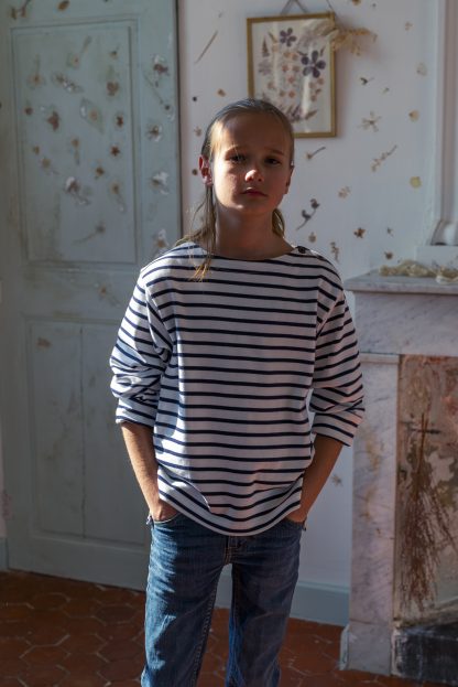 Child wearing the Pénélope Marinière sewing pattern from Ready to Sew on The Fold Line. A top pattern made in cotton jersey or thick double-knit fabrics, featuring a straight silhouette, boat neckline, two button shoulder closure, full length sleeves, side vents and a slight high low hem.