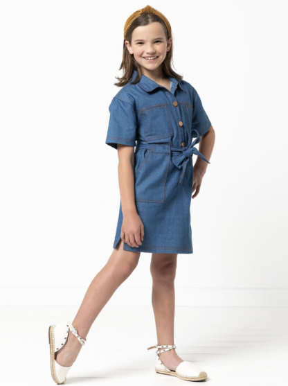 Child wearing the Child/Teen Demi Dress sewing pattern from Style Arc on The Fold Line. A dress pattern made in denim, cotton drill or heavy linen fabrics, featuring collar and stand, short sleeves, front button tab, waist tie, chest patch pockets, skirt pockets, and side hem splits.