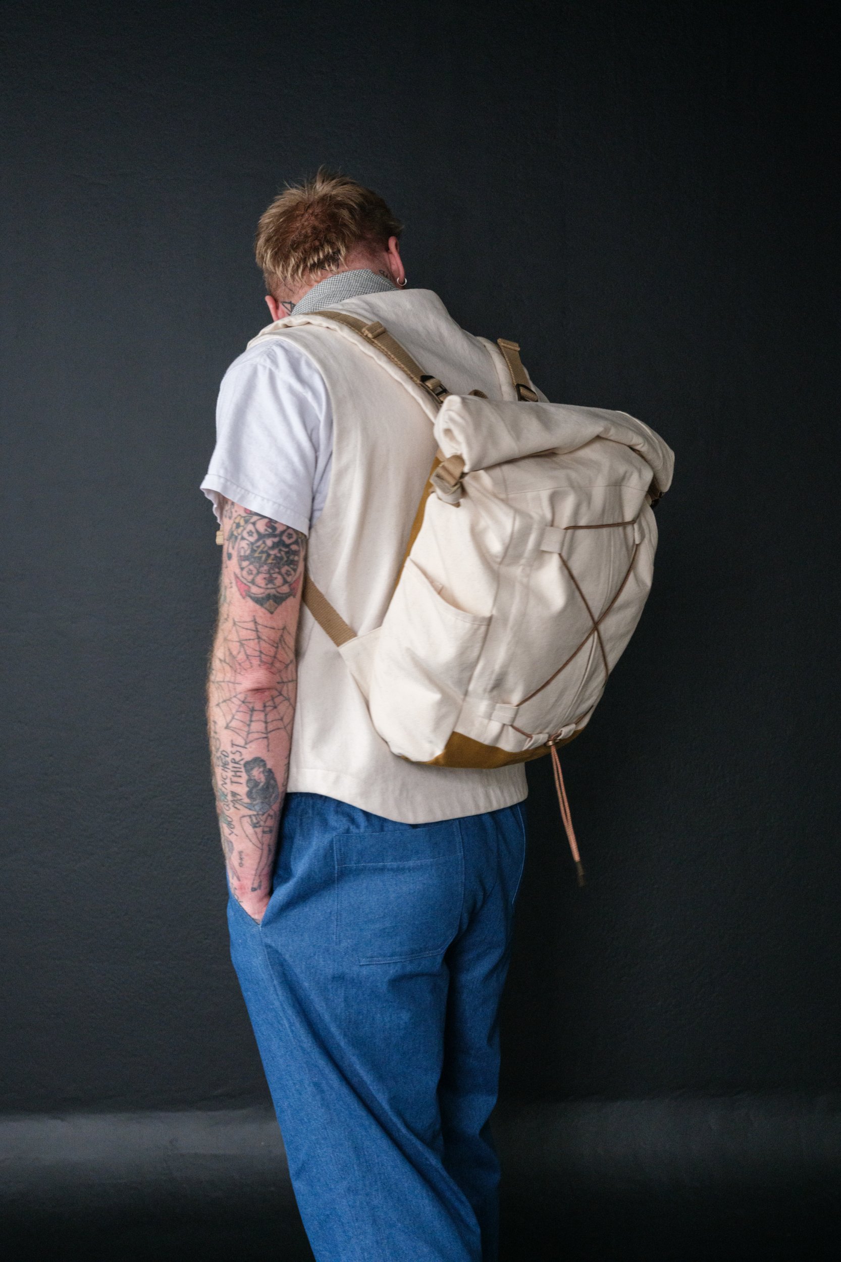 Man wearing the Francli Day Pack sewing pattern from Merchant & Mills on The Fold Line. A back pack pattern made in oilskin, dry oilskin, cotton canvas, drill, or mid weight denim fabrics, featuring water bottle pockets, internal laptop pocket, adjustable shoulder straps, and fold-over top.