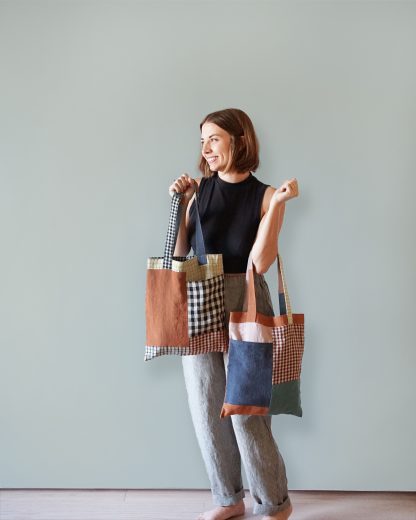 Woman holding the Stash Pocket Totes sewing pattern from Matchy Matchy Sewing Club on The Fold Line. A tote bag pattern made in medium weight cotton, woven, twill, and canvas fabrics, featuring a full lining, outside pocket and shoulder straps.