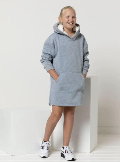 Child wearing the Child/Teen Zara Hooded Dress sewing pattern from Style Arc on The Fold Line. A knit dress pattern made in Sweater knit or French terry fabrics, featuring a lined hood, sleeves with cuffs, centre front pocket, and side splits finished with tape.