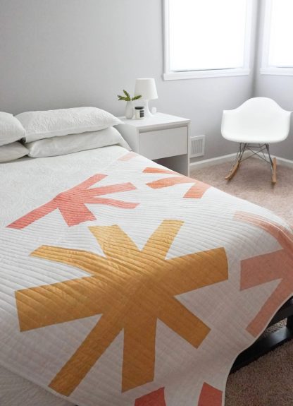 Photo showing the Asterisks Quilt sewing pattern from Modern Handcraft on The Fold Line. A quilt pattern made in quilting cotton fabrics, featuring a white asterisk design on a multi coloured background, using square and triangular shapes.