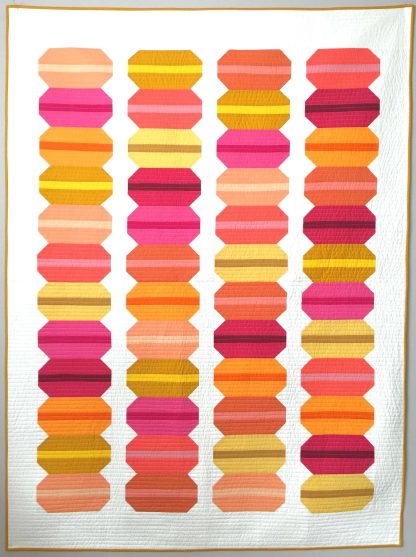 Photo showing the French Macaron Quilt sewing pattern from Modern Handcraft on The Fold Line. A quilt pattern made in quilting cotton fabrics, featuring stacks of macarons in colourful reds, pinks and oranges on a cream background.