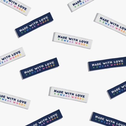 Photo showing 'Made With Love and Swear Words' Cotton Labels from Kylie & The Machine on The Fold Line. A washable, durable, and non-scratchy cotton label featuring a cream or navy background with bold navy or cream coloured text and rainbow mini text, they are all ready to be sewn into your handmade clothes.