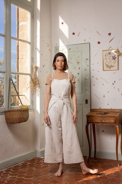 Woman wearing the Pablo Overall sewing pattern from Ready To Sew on The Fold Line. An overall/apron combo pattern made in linen, or ramie, fabrics, featuring a wrap around the hips and waist feature, relaxed fit, two front and four back darts, wide legs, cropped length, narrow shoulder straps that cross at the back and tie at the waist, front and back patch pockets,