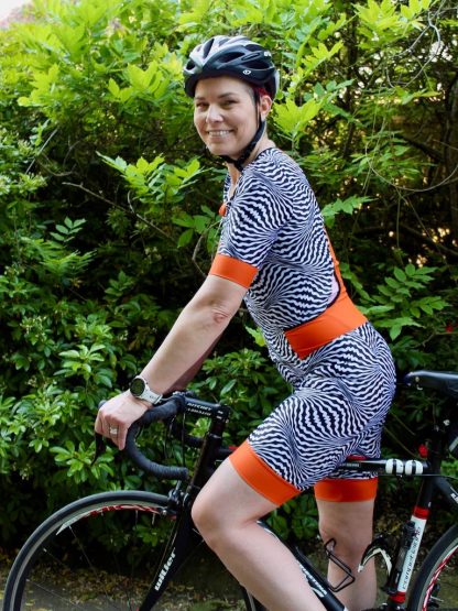 Woman wearing the Triumph Suit sewing pattern from Fehr Trade on The Fold Line. An all-in-one cycling suit pattern made in lycra fabric, featuring short sleeves, no side seams, front and back princess seams, banded shorts hem, front zip fastener, four back pockets, and two easy access side pockets.