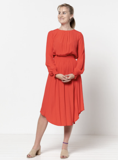 Woman wearing the Asha Dress sewing pattern from Style Arc on The Fold Line. A dress pattern made in silk, rayon or georgette fabrics, featuring a midi length, relaxed fit, slight blouson bodice, shirred waistline, gathered skirt, long sleeve with shirred cuff, round neck with front gathers, back neck button and loop closure, and high-low hem.